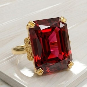 Puede incluir: Un anillo de oro con una gran gema roja oscura, rectangular y facetada. La gema está engastada en un marco de oro con detalles decorativos en cada esquina. La banda del anillo también es de oro, con intrincados detalles de filigrana.