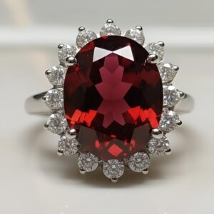 Pode incluir: Um anel de prata com uma grande pedra preciosa vermelha escura de corte oval, rodeada por um halo de pedras brancas redondas menores. O design do anel é elegante e adequado para ocasiões especiais.