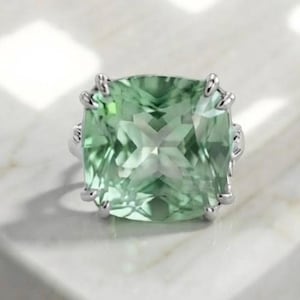 May include: A silver ring featuring a large, square-cut, light green gemstone with faceted surfaces. The stone is held in place by four silver prongs at each corner, creating a clear, geometric display of the pale green, translucent mineral.