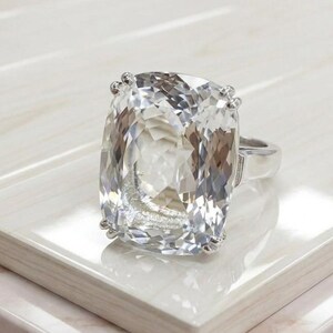 May include: A silver-toned ring featuring a large, rectangular cushion-cut clear gemstone with faceted surfaces. The stone is secured by four prongs at the corners, set upon a smooth, polished metal band that tapers toward the back of the ring.