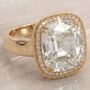 Könnte beinhalten: Ein goldener Ring mit einem großen, quadratisch geschliffenen, klaren Edelstein. Der Edelstein ist von einem Kranz aus kleineren, klaren Steinen und einem gedrehten Goldseildesign umgeben. Der Ringband ist dick und poliert.