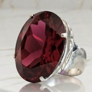 Puede incluir: Un llamativo anillo con una gran gema roja oscura de forma ovalada engastada en una banda de plata. La gema está facetada, reflejando la luz. El anillo tiene un diseño audaz. El engaste incluye pequeñas garras.