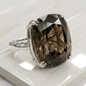 Puede incluir: Un anillo de plata con una gran gema de cuarzo ahumado de talla cojín rectangular. La gema marrón está sujeta por garras. La banda del anillo es plateada y las facetas de la piedra crean un efecto brillante al reflejar la luz.