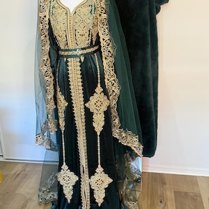 May include: A dark green velvet dress with gold embroidery and a matching veil. The dress features intricate gold patterns along the neckline, sleeves, and hem. A gold belt cinches the waist. The veil is sheer with gold trim.
