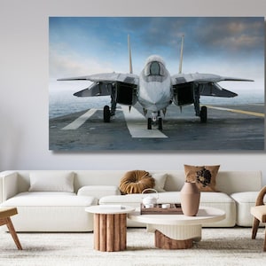 May include: A rectangular wall art print featuring a front-facing gray fighter jet positioned on the flight deck of an aircraft carrier. The aircraft is centered against a backdrop of a cloudy blue sky and the open ocean, with visible landing markings.