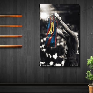 Puede incluir: Un cuadro rectangular de pared que presenta una fotografía en blanco y negro de un futbolista sosteniendo una camiseta azul y roja con el número 10 y el nombre Messi impresos en amarillo en la parte posterior, sobre una pared de madera oscura.