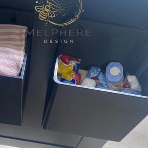 Könnte beinhalten: Schwarzes Aufbewahrungsregal mit zwei Fächern. Ein Fach enthält ein gefaltetes rot-weiß gestreiftes Handtuch. Das andere Fach enthält verschiedene bunte Waschmittel-Pods und -Tabletten. Der Text "MELPHERE DESIGN" ist sichtbar.