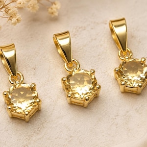 Puede incluir: Tres collares con colgantes dorados con gemas hexagonales de color amarillo claro. Los colgantes tienen un diseño sencillo con un lazo para una cadena. Las gemas están facetadas. El fondo es de color neutro.