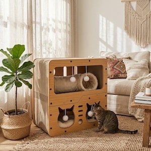 Peut inclure: Une maison pour chat en bois avec un tunnel et deux ouvertures en forme de chat. La maison pour chat a une palette de couleurs neutres et comprend des pompons suspendus pour jouer. Un chat tigré est assis près de la maison. Un pot de plante et un tapis tissé sont également dans l'image.