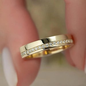 May include: A gold band ring with a row of small, clear gemstones set into the band. The ring is held between fingers, and the gold band has a polished, reflective surface. The ring is a classic design, suitable for various occasions.