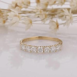 May include: A gold band ring featuring a series of square-cut, clear gemstones. The ring is set against a soft, blurred background with white flowers. The ring is a piece of jewelry.