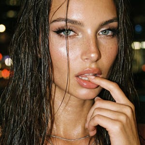 May include: A close-up portrait of a person with wet, dark brown hair and light skin. The person has blue-green eyes, freckles, and is wearing a silver necklace. The background is blurred with bokeh lights.