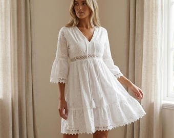Könnte beinhalten: Weißes Eyelet-Kleid mit V-Ausschnitt, Glockenärmeln und einem Stufenrock. Das Kleid hat aufwendige Ausschnitte und eine Quastenbindung am Ausschnitt. Das knielange Kleid scheint aus leichtem Stoff gefertigt zu sein.