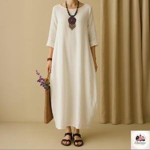 Op de afbeelding: Een witte linnen jurk met een ronde hals en driekwart mouwen. De jurk wordt gecombineerd met bruine sandalen en een geweven draagtas met bloemen. Een kleurrijke ketting voegt een decoratief tintje toe. De jurk heeft een relaxte, vloeiende silhouet.