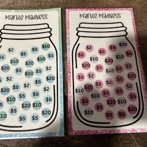 May include: Two "Marble Madness" savings tracker cards. One has a blue and green border with blue circles, the other has a pink and purple border with pink circles. Each card features a jar graphic filled with dollar amounts.