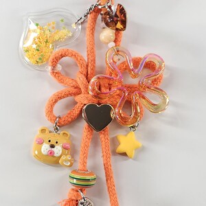 May include: Orange keychain with various charms. Features a clear heart-shaped container with yellow star confetti, a bear charm, a flower charm, a heart charm, and a yellow star charm. The keychain has a lobster clasp.