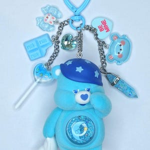Op de afbeelding: Een blauwe Care Bear sleutelhanger met een hartvormige sluiting. De sleutelhanger heeft een pluchen beer met een maan- en sterrenontwerp, omringd door bedels, waaronder een hart, een strik, een melkpak en een 'Be Mine' hart.