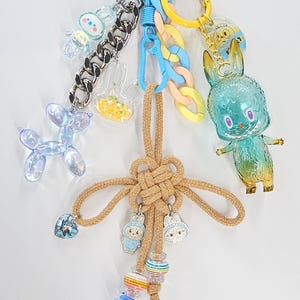 May include: A multi-layered keychain featuring a tan knotted rope, a large teal and yellow gradient rabbit figure, a blue balloon dog charm, clear acrylic charms with stars, and colorful plastic chain links attached to silver and blue metal rings.