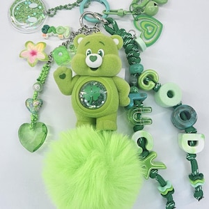 Op de afbeelding: Een groene sleutelhanger met een gevlokt berenfiguur met een klaver op de buik, een grote limoengroene imitatiebont pompon en diverse bedels waaronder een smiley, een bloem en kralen die de woorden GOOD LUCK in het groen spellen.