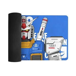 May include: A blue and white rectangular mousepad featuring a detailed illustration of a Gundam robot. The robot has red, white, yellow, and blue accents. The mousepad is rolled up on the left side, revealing a black textured underside.
