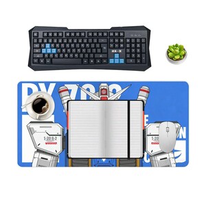 May include: A black gaming keyboard with blue keys sits above a blue mousepad featuring a robot design. A white mouse, a cup of coffee, a notebook, and a small succulent plant are also visible. The mousepad has the text "DV 700".