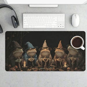 May include: A rectangular desk mat featuring an illustration of five frogs wearing pointed wizard hats and robes, standing in a dark forest setting with small lit candles and mushrooms on the ground, with a white coffee mug placed on the right side.