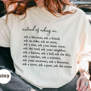 May include: An ivory t-shirt featuring black text that reads: instead of asking ai: ask a librarian, ask a friend, ask an elder, ask an artist, ask a tree, ask your inner voice, ask the void, ask your neighbor, ask a barista, ask a kid, ask the sky.
