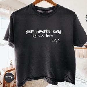 May include: A charcoal gray short-sleeve t-shirt featuring white printed text that reads your favorite song lyrics here -artist. The shirt is displayed on a wooden hanger and has a crew neckline with a standard fit and a solid color finish.