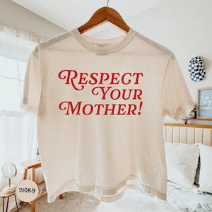 May include: An ivory short-sleeved t-shirt featuring the red text "RESPECT YOUR MOTHER!" printed in a bold, stylized serif font across the chest. The garment has a classic crew neckline and is displayed on a wooden hanger.
