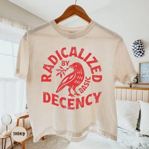 May include: An ivory short-sleeve t-shirt featuring a red graphic print of a bird holding a branch. The text "RADICALIZED BY BASIC DECENCY" is printed in a circular pattern around the bird in a distressed, weathered red font style.