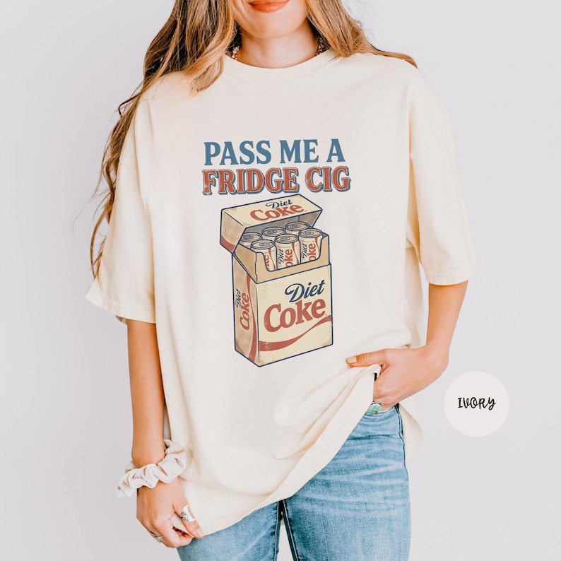 Może przedstawiać: T-shirt z kr&oacute;tkim rękawem w kolorze kości słoniowej z nadrukiem graficznym przedstawiającym opakowanie puszek Diet Coke. Tekst nad grafiką brzmi "PASS ME A FRIDGE CIG" niebieskimi i czerwonymi literami. Wz&oacute;r przedstawia kartonowe pudełko z puszkami.