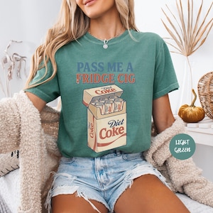Może przedstawiać: Zielony t-shirt z napisem "PASS ME A FRIDGE CIG" niebieskimi i pomarańczowymi literami nad grafiką przedstawiającą otwarte pudełko puszek Diet Coke. Wz&oacute;r zawiera logo Diet Coke na pudełku oraz na poszczeg&oacute;lnych puszkach napoju wewnątrz.