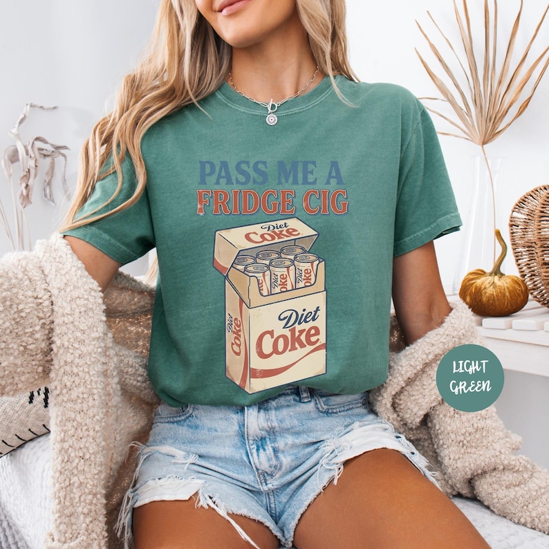 Może przedstawiać: Zielony t-shirt z napisem "PASS ME A FRIDGE CIG" niebieskimi i pomarańczowymi literami nad grafiką przedstawiającą otwarte pudełko puszek Diet Coke. Wz&oacute;r zawiera logo Diet Coke na pudełku oraz na poszczeg&oacute;lnych puszkach napoju wewnątrz.