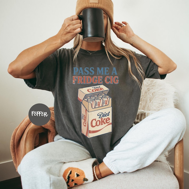 Może przedstawiać: Ciemnoszary t-shirt z kr&oacute;tkim rękawem z napisem "PASS ME A FRIDGE CIG" niebieskimi i czerwonymi literami nad grafiką przedstawiającą otwarte kartonowe pudełko zawierające sześć srebrnych puszek napoju "Diet Coke" z czerwonym logo.