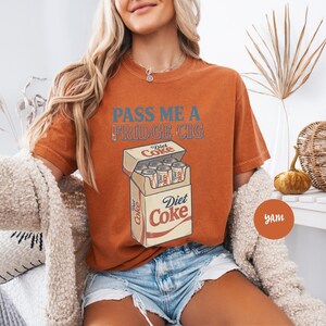 Może przedstawiać: T-shirt z kr&oacute;tkim rękawem w kolorze przypalonej pomarańczy z niebieskim napisem "PASS ME A FRIDGE CIG" oraz grafiką otwartej paczki papieros&oacute;w wypełnionej puszkami Diet Coke, z logo Diet Coke nadrukowanym na przodzie opakowania.