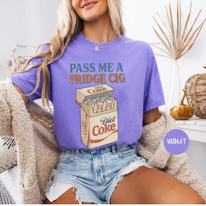 Może przedstawiać: Fioletowy t-shirt z kr&oacute;tkim rękawem z napisem PASS ME A FRIDGE CIG zielonymi i pomarańczowymi literami nad grafiką przedstawiającą otwarty karton wypełniony puszkami Diet Coke. Koszulka ma okrągły dekolt i luźny kr&oacute;j z nadrukiem w stylu vintage.