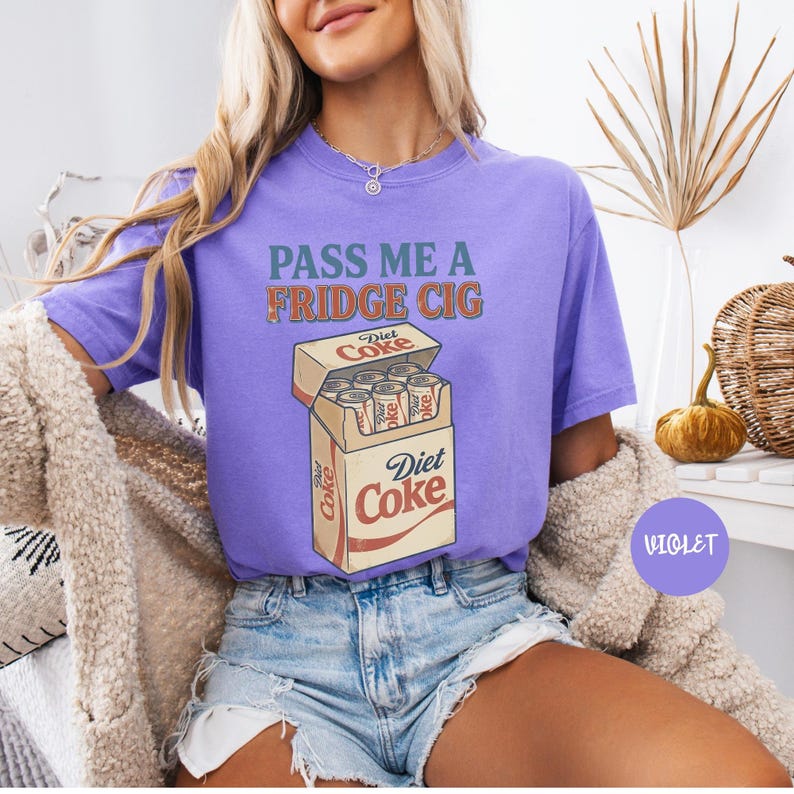 Może przedstawiać: Fioletowy t-shirt z kr&oacute;tkim rękawem z napisem PASS ME A FRIDGE CIG zielonymi i pomarańczowymi literami nad grafiką przedstawiającą otwarty karton wypełniony puszkami Diet Coke. Koszulka ma okrągły dekolt i luźny kr&oacute;j z nadrukiem w stylu vintage.