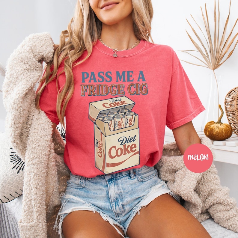 Może przedstawiać: Koralowy t-shirt z kr&oacute;tkim rękawem z napisem "PASS ME A FRIDGE CIG" niebieskimi i brązowymi literami nad grafiką przedstawiającą otwarty karton wypełniony srebrnymi puszkami Diet Coke oraz logo Diet Coke z przodu.