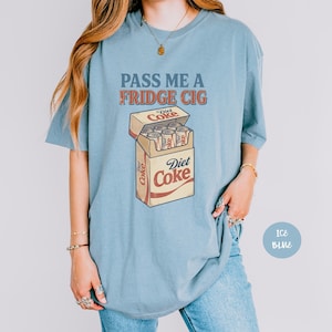 Może przedstawiać: Jasnoniebieski t-shirt z grafiką przedstawiającą paczkę papieros&oacute;w wypełnioną puszkami Diet Coke. Tekst nad grafiką brzmi PASS ME A FRIDGE CIG w postarzanym stylu. Logo Diet Coke jest nadrukowane na przedniej części paczki papieros&oacute;w.