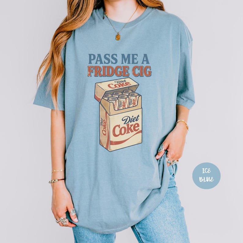 Może przedstawiać: Jasnoniebieski t-shirt z grafiką przedstawiającą paczkę papieros&oacute;w wypełnioną puszkami Diet Coke. Tekst nad grafiką brzmi PASS ME A FRIDGE CIG w postarzanym stylu. Logo Diet Coke jest nadrukowane na przedniej części paczki papieros&oacute;w.