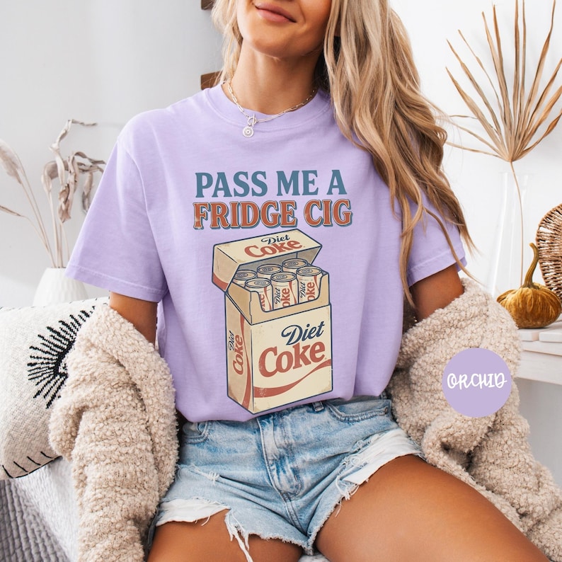 Może przedstawiać: Jasnofioletowy t-shirt z napisem "PASS ME A FRIDGE CIG" niebieskimi i pomarańczowymi literami nad grafiką przedstawiającą otwarte kartonowe pudełko wypełnione srebrnymi puszkami Diet Coke, z nazwą marki nadrukowaną na przodzie pudełka.