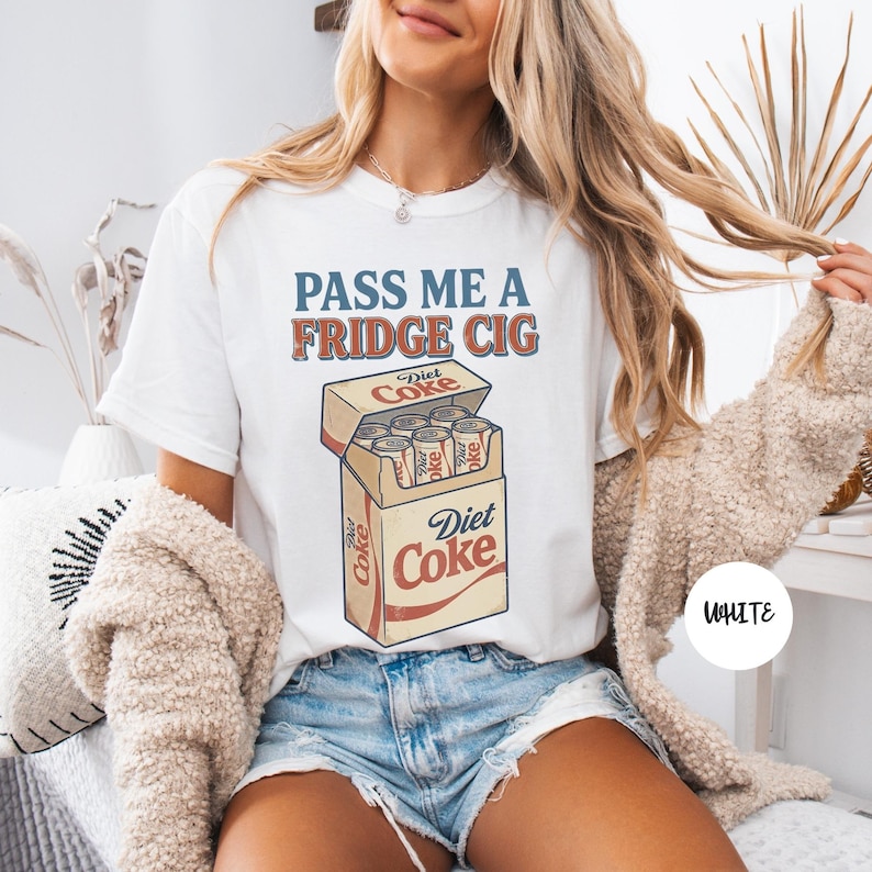 Może przedstawiać: Biały t-shirt z napisem PASS ME A FRIDGE CIG niebieskimi i pomarańczowymi literami nad grafiką przedstawiającą otwarty karton wypełniony puszkami Diet Coke. Projekt zawiera logo Diet Coke w kolorze czerwonym i białym na kartonie.