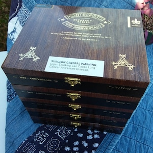 May include: A stack of five wooden cigar boxes with gold hinges and labels. The top box is labeled "Montecristo Nicaragua Series 1935 Anniversary" and has a warning label that reads "Surgeon General Warning: Cigar Smoking Can Cause Lung Cancer And Heart Disease."
