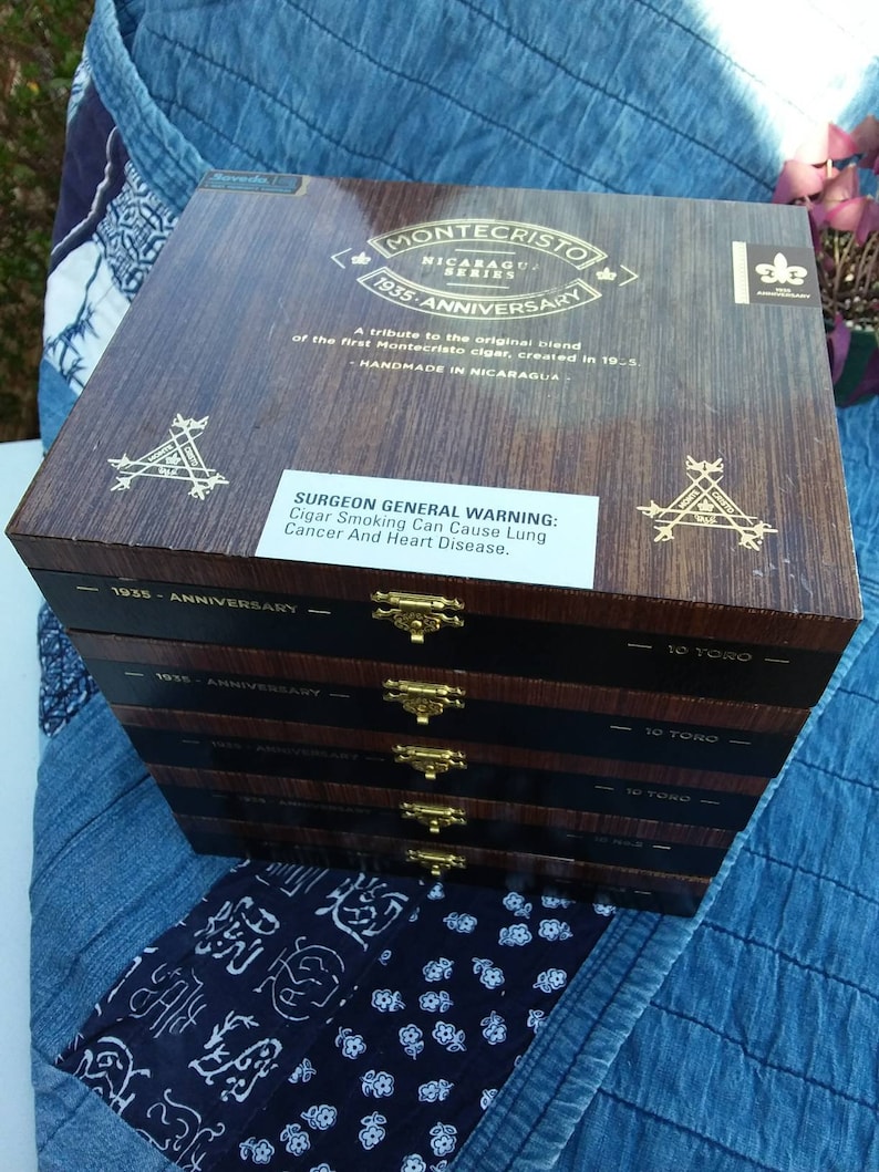 May include: A stack of five wooden cigar boxes with gold hinges and labels. The top box is labeled "Montecristo Nicaragua Series 1935 Anniversary" and has a warning label that reads "Surgeon General Warning: Cigar Smoking Can Cause Lung Cancer And Heart Disease."