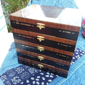 May include: A stack of six wooden cigar boxes with black lids. Each box has a gold latch and a label with the text "1935 - Anniversary" and "10 Toro" or "10 No.2".