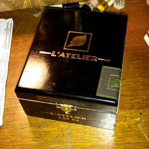May include: A black wooden box with a gold leaf design and the text "L'Atelier Lat 52". The box has a gold leaf design of a leaf and the text "L'Atelier" on the top.