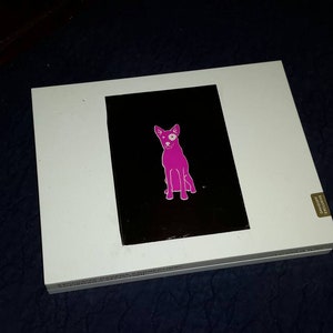 May include: A white box with a black and white image of a pink dog with one white eye on the front. The box has a gold sticker on the side.