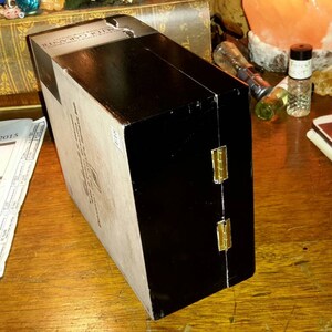 May include: A black wooden box with gold hinges. The box has a white label with black text. The box is closed and sitting on a wooden surface.