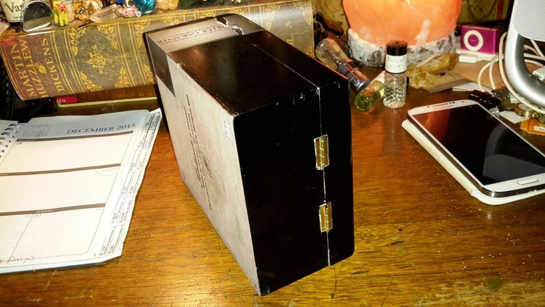 May include: A black wooden box with gold hinges. The box has a white label with black text. The box is closed and sitting on a wooden surface.