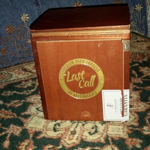 May include: A wooden box with a gold label that says "Premium Aged Tobacco Last Call by AJ Fernandez".